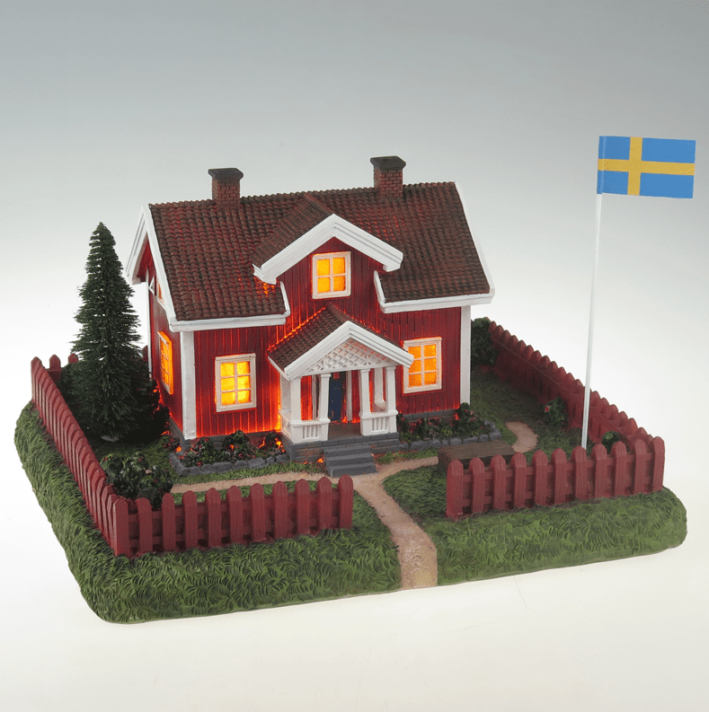 Sommarby ROUGE De Pixie Design - "Le Cottage Rouge en Été" - est une lampe décorative en céramique représentant une maison d'été suédoise classique avec un jardin environnant et un mât de drapeau.