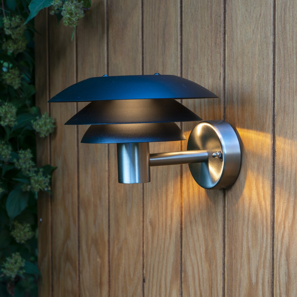 DL20 Acier inoxydable lampe d’extérieur De Dyberg Larsen - La lampe est dotée de trois abat-jour emblématiques de différentes tailles qui diffusent une lumière magique et est fabriquée en acier inoxydable avec d'élégants abat-jour noirs mats.