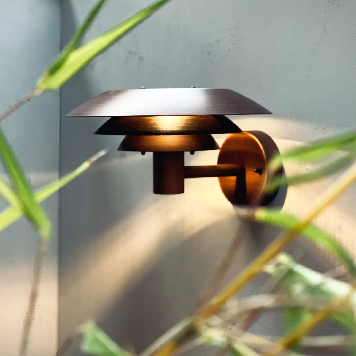 DL25 Cuivre lampe d’extérieur De Dyberg Larsen - Med sina tre ikoniska lampsk&auml;rmar sprider den ett fascinerande ljus, idealiskt f&ouml;r att skapa en st&auml;mningsfull atmosf&auml;r l&auml;ngs fasaden eller i andra utomhusmilj&ouml;er.