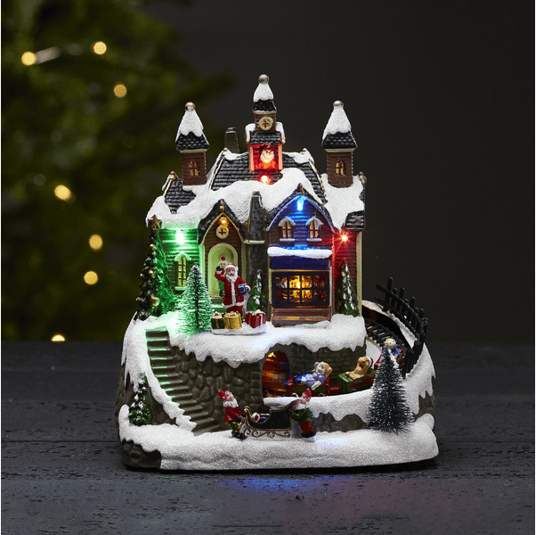 Trainville Multicolore De Star Trading - Village de Noël décoratif composé de maisons avec des tours, de fenêtres lumineuses et bruyantes, et d'un Père Noël debout qui fait signe de la main.